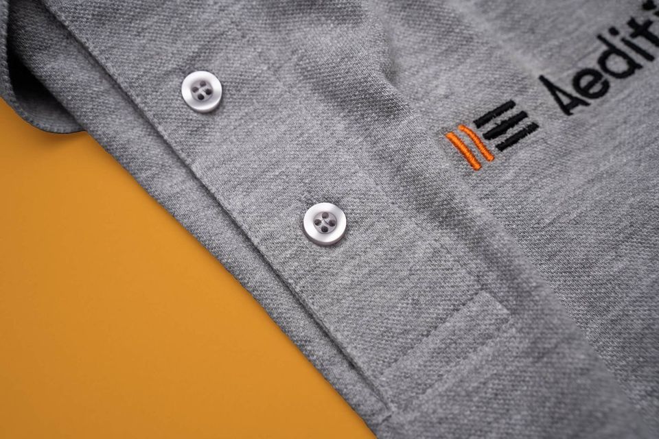 Graues Poloshirt mit gesticktem Firmenlogo auf orangem Untergrund